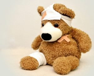 Brown teddy bear with a bandage on his head, bandage on his leg, and crossed band aids on his chest.